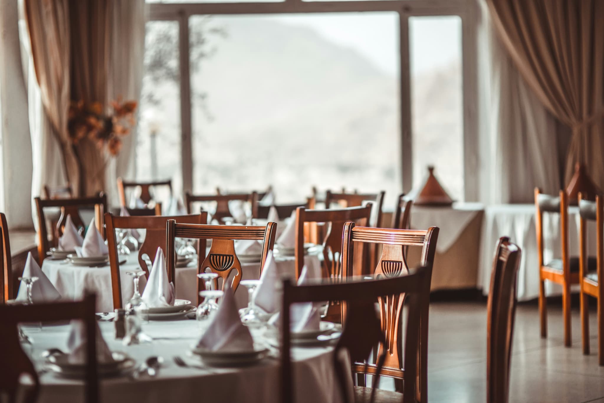 An elegant dining room is set with neatly folded napkins, glassware, and wooden chairs beside large windows with soft curtains.