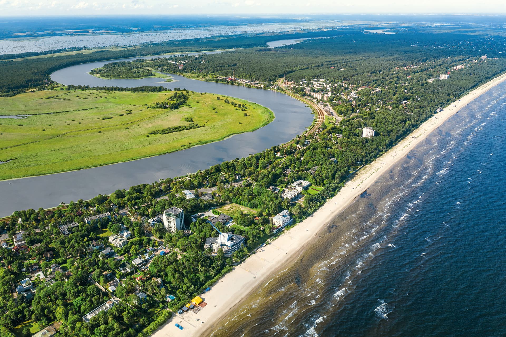 An aerial view shows a long sandy beach beside a forested coastal town, with a winding river and wide green marshland inland.