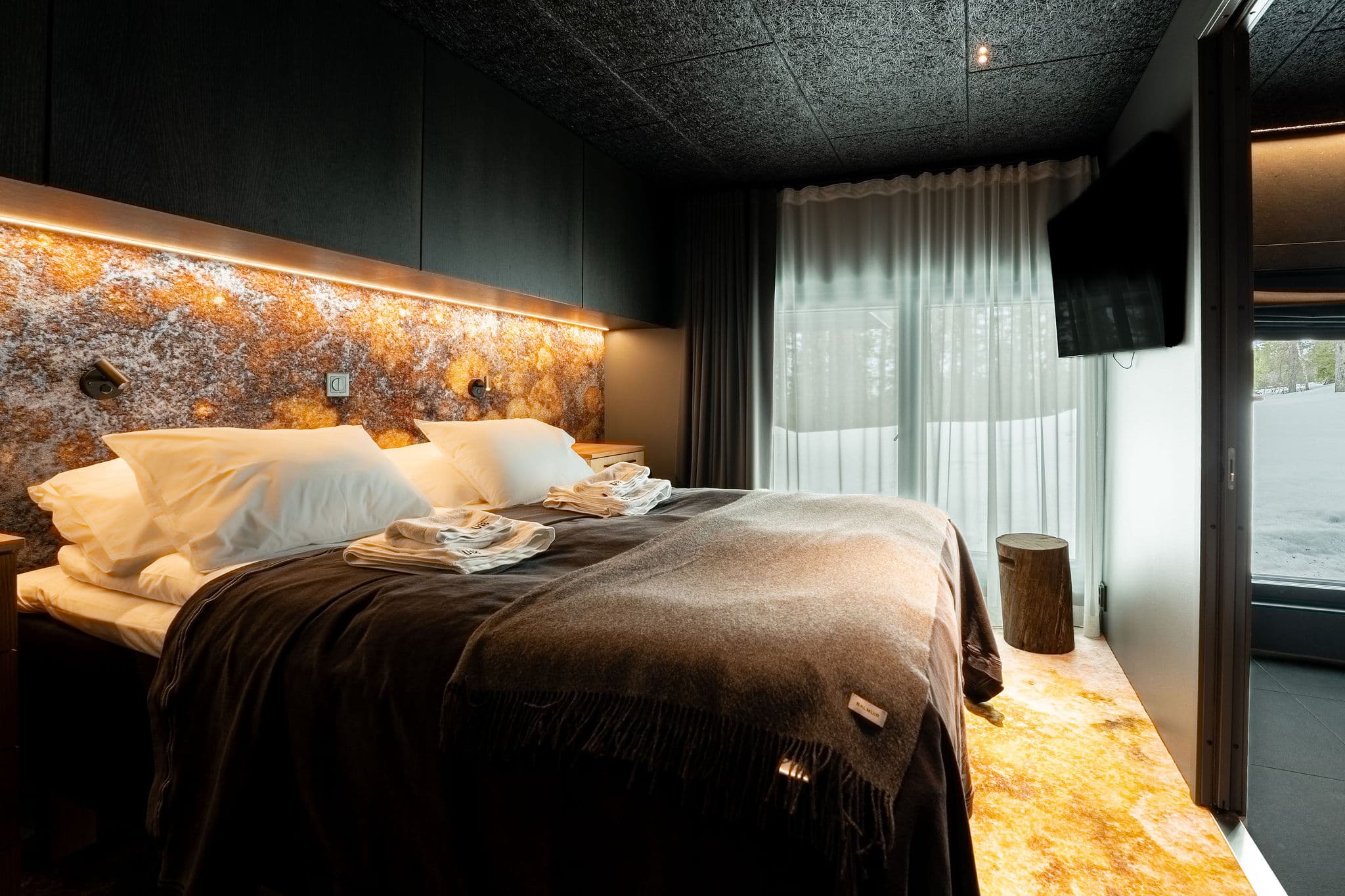 Luxury chalet bedroom interior with a bed, textured stone wall, warm lighting, and a view toward a snow-covered patio through open glass doors.