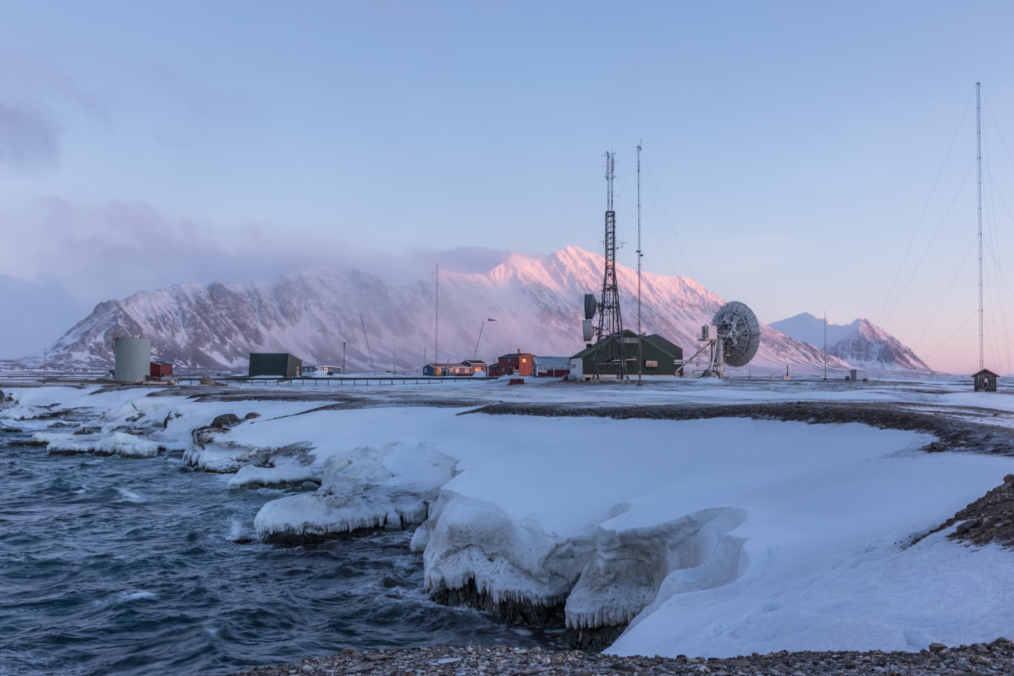 Snow-covered mountains rise behind a coastal radio station with tall antenna towers and icy shoreline water at dusk.