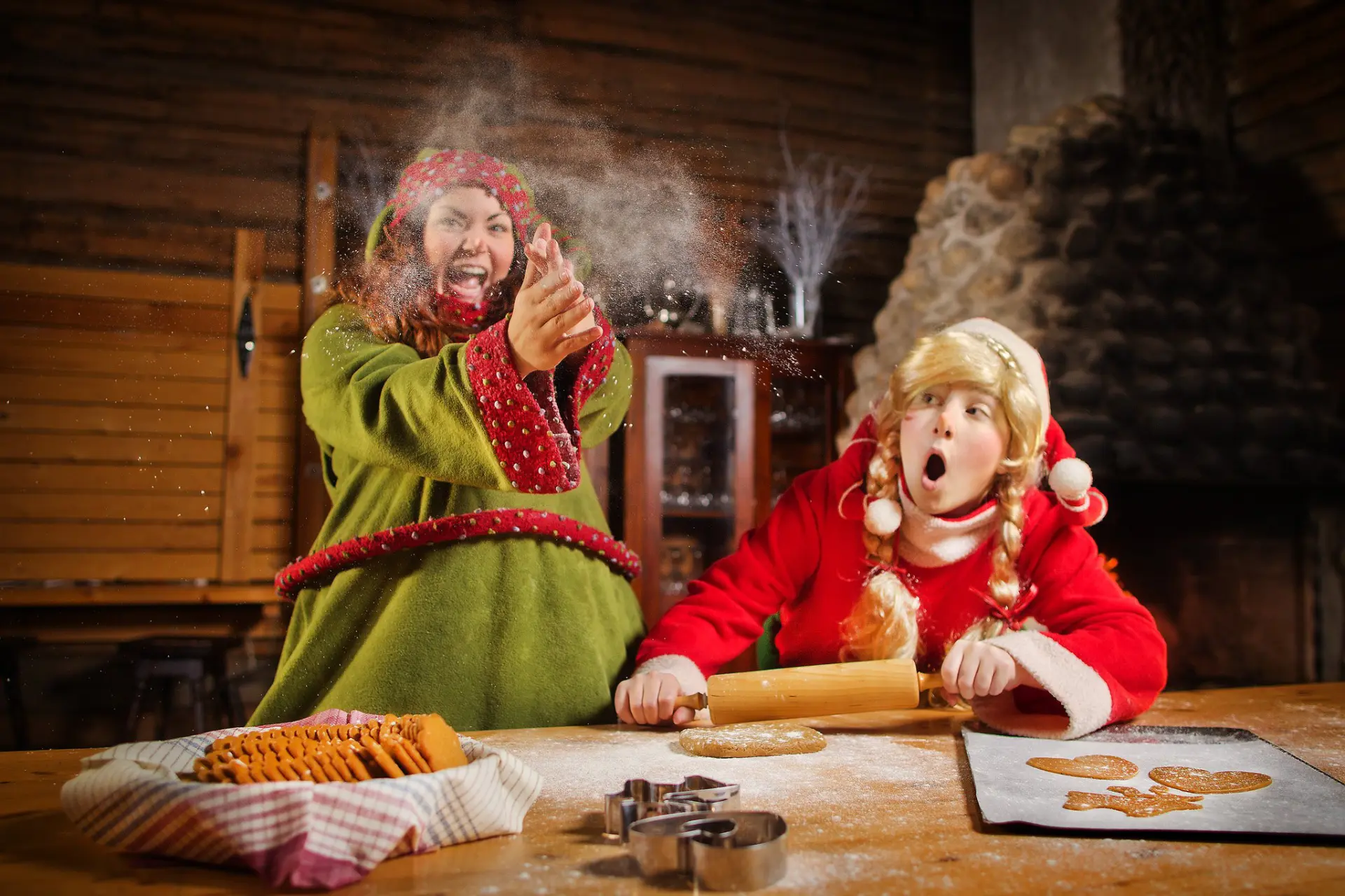 Two people in festive elf costumes bake gingerbread at a flour-dusted table inside a rustic wooden room.