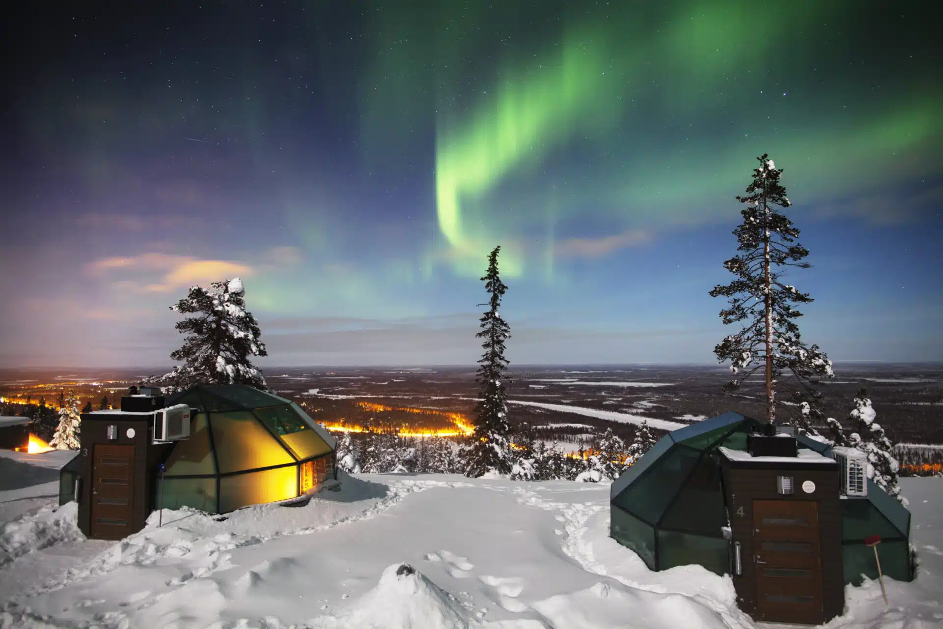 Snow-covered glass igloo cabins glow on a winter hillside beneath a vivid green aurora in a starry night sky.