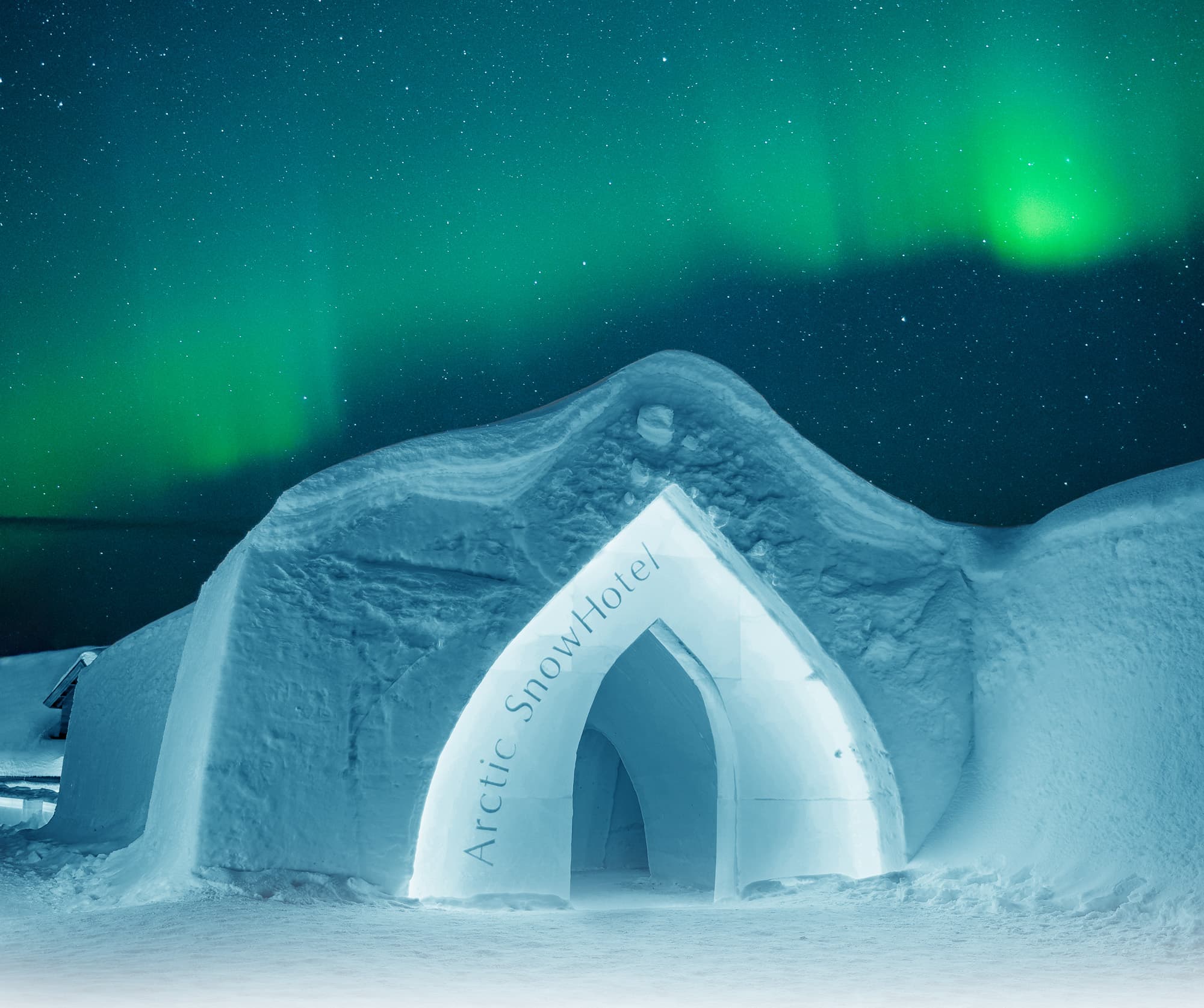 Snow-covered arctic ice hotel entrance with arched doorway beneath green aurora lights in the night sky.