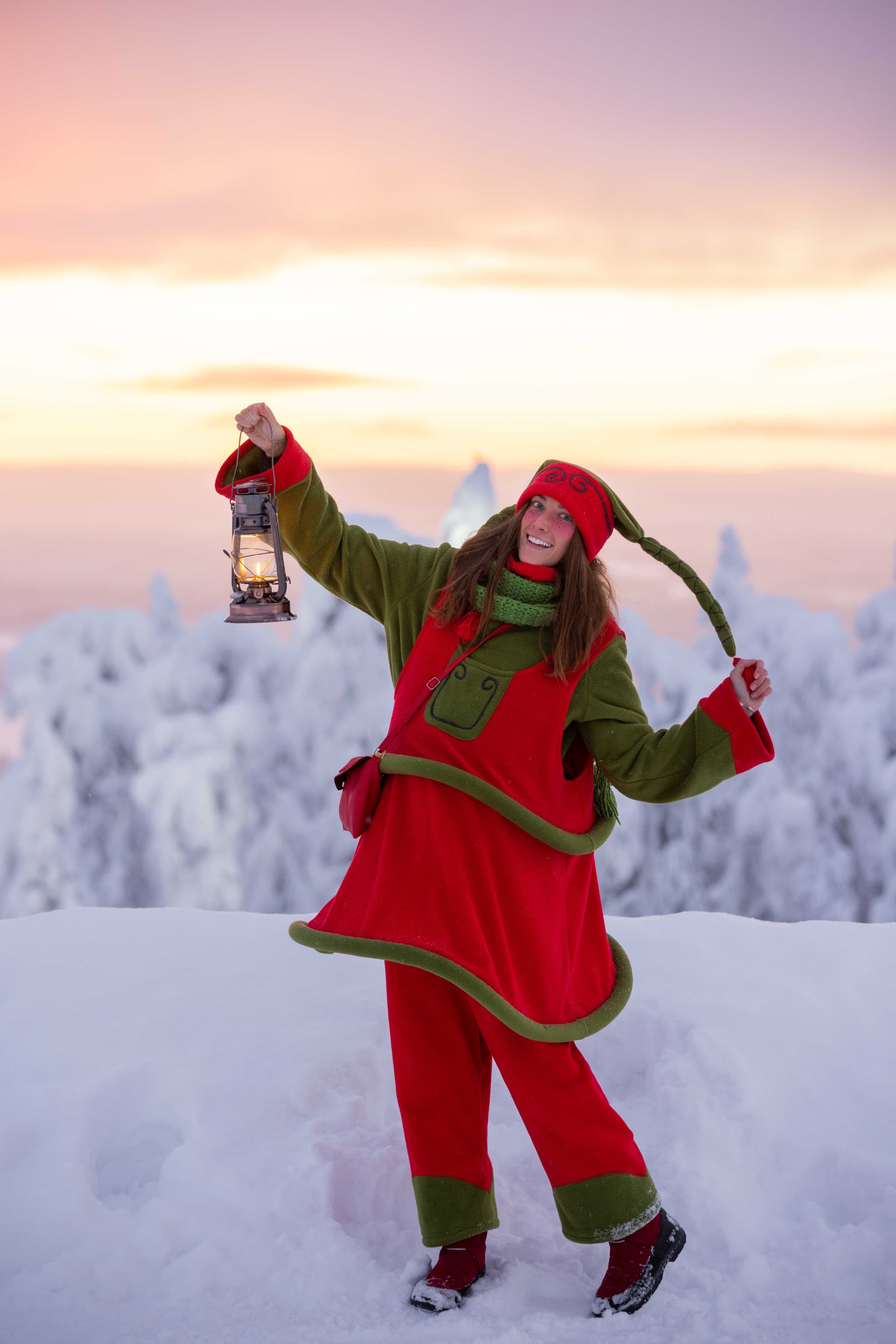A smiling woman in a red-and-green winter outfit holds up a lit lantern while standing in deep snow at sunset.