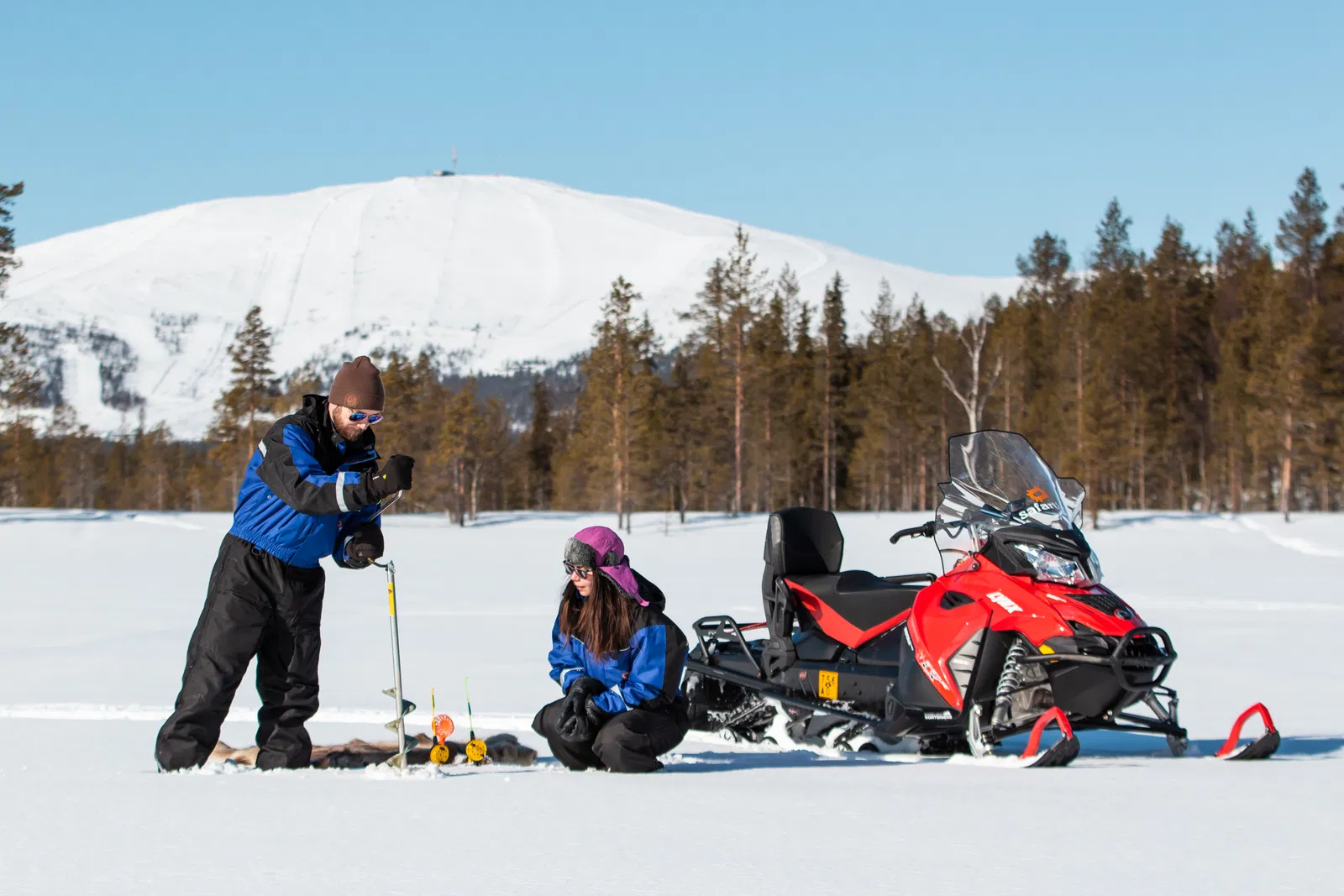 Two people ice fish on a frozen lake beside a red snowmobile with a snowy mountain and pine forest in the background.
