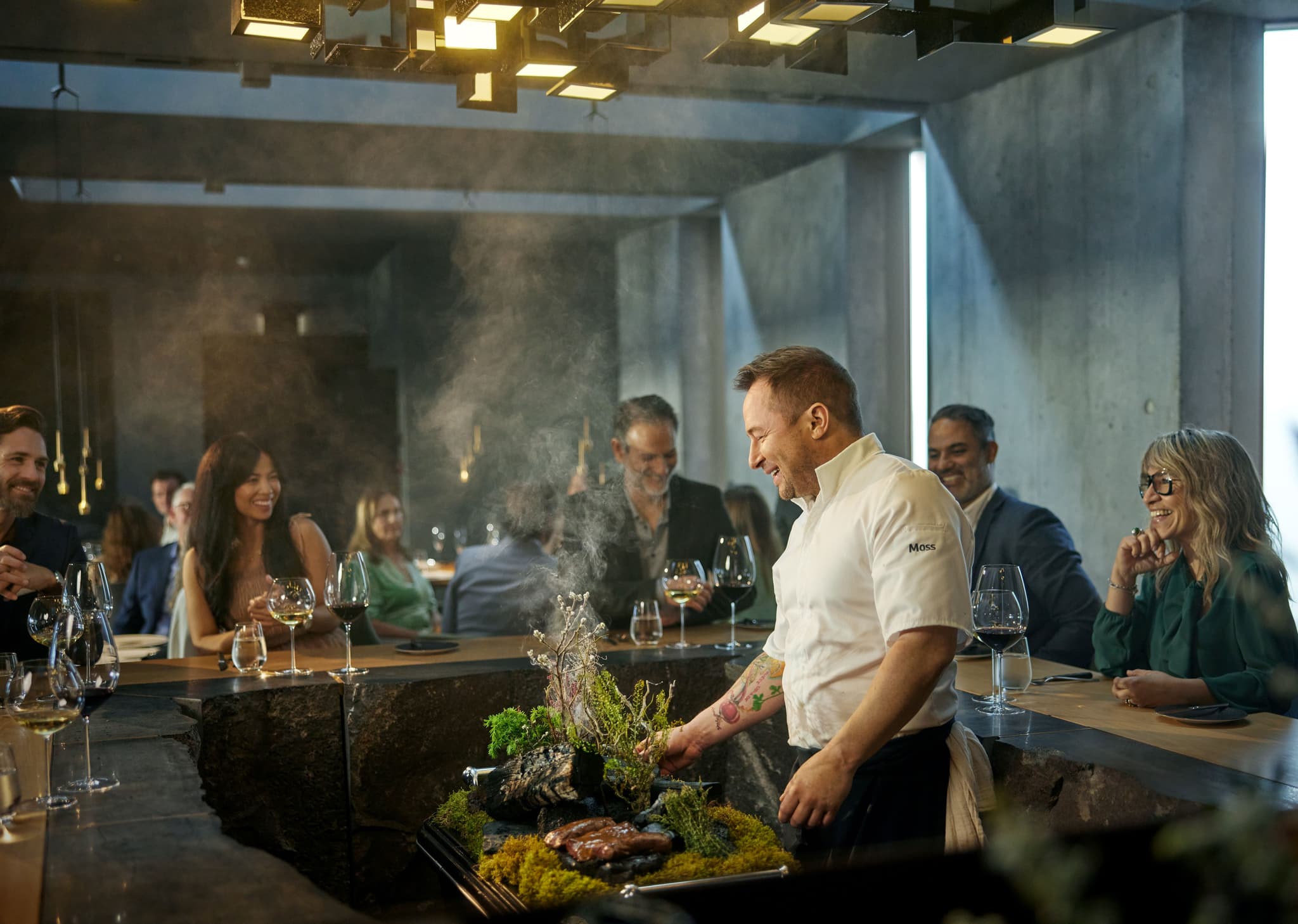 Chef in white jacket prepares food at a restaurant table as diners sit with wine glasses and candles; light steam rises from the dish.