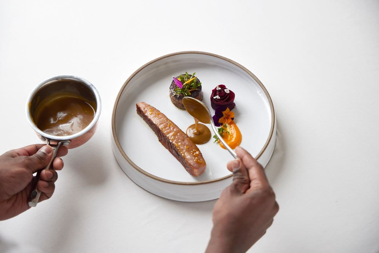 A plated gourmet dish is being finished with sauce from a small metal pot while a spoon is held above the plate.