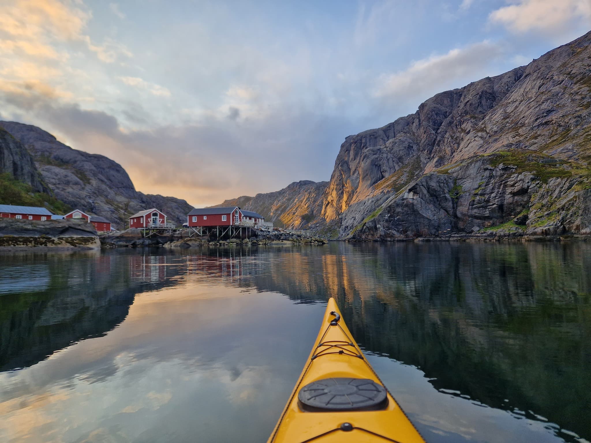 Yellow kayak in calm fjord waters at dusk with red cabins on stilts and steep rocky mountains under cloudy skies.