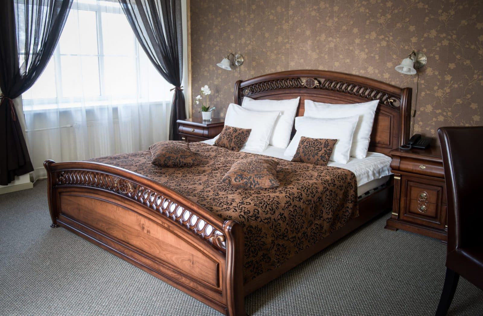 A warmly decorated bedroom features a carved wooden bed dressed in patterned brown bedding, white pillows, and matching bedside tables beside a bright window with dark curtains.